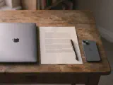 A small wooden desk at dawn — a white ceramic coffee mug, a closed silver laptop, a printed morning brief, and a phone face-down, lit by warm low-angle sunlight.