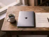 A small wooden desk at dawn — a white ceramic coffee mug, a closed silver laptop, a printed morning brief, and a phone face-down, lit by warm low-angle sunlight.