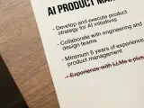 A printed AI Product Manager job description on a wooden desk, its bottom bullet (experience with LLMs a plus) struck through in red pen, with a handwritten margin note reading make it falsifiable.